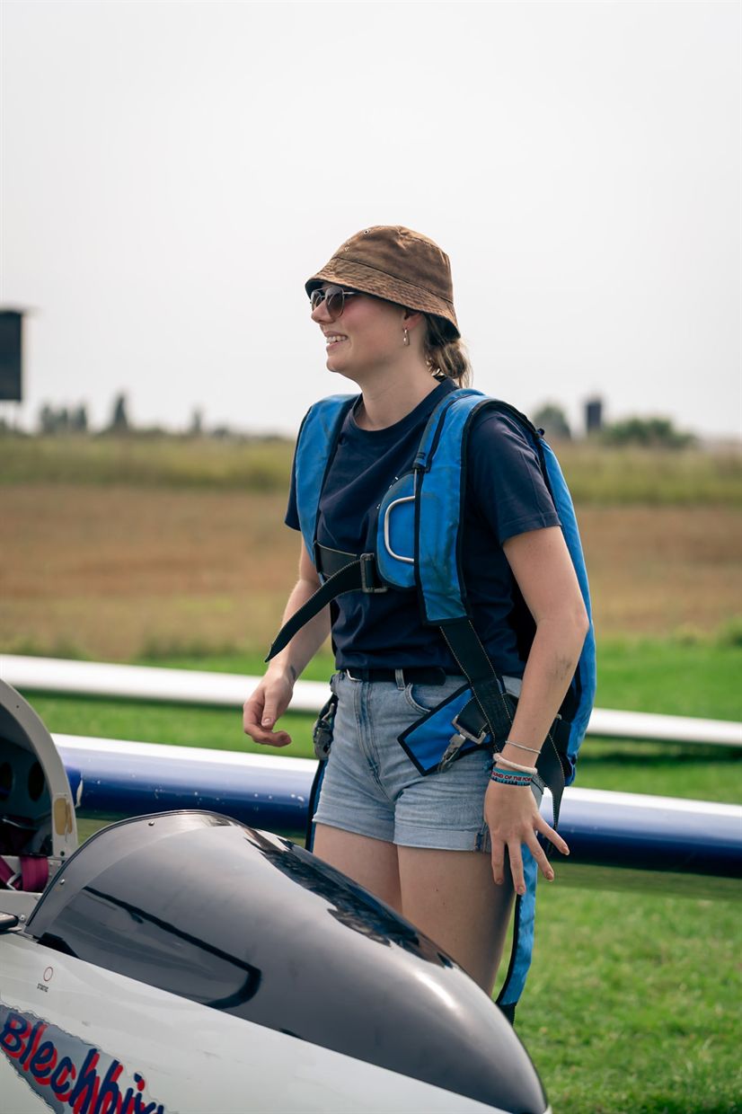 Frau mit Hut und Fallschirmrucksack lacht neben einem Segelflugzeug